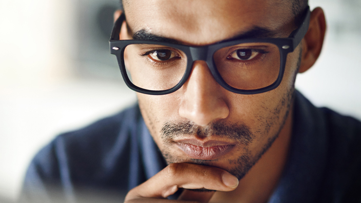 A man with a contemplative expression looks at a computer monitor in an office setting