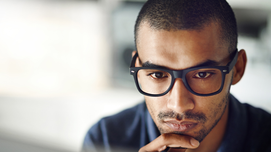 A man with a contemplative expression looks at a computer monitor in an office setting