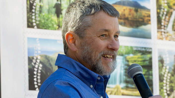 A man stands at a lectern in a forested setting, near a poster displaying natural scenery stamps