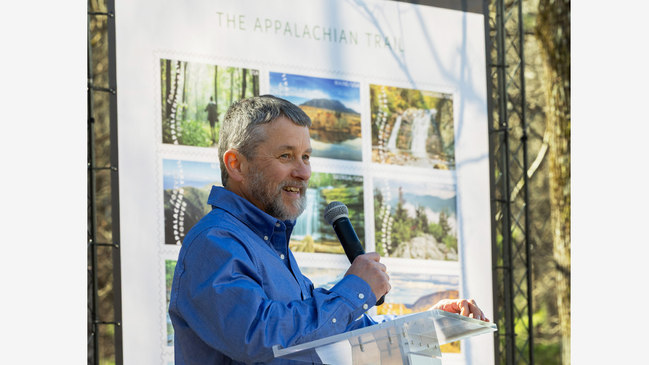 A man stands at a lectern in a forested setting, near a poster displaying natural scenery stamps