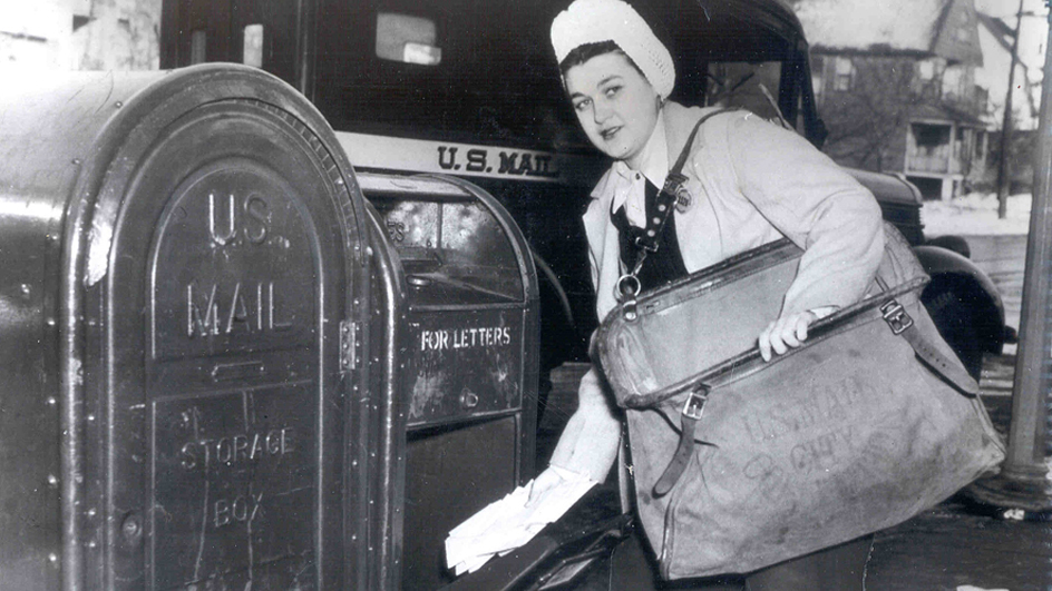 A black-and-white photo of a woman wearing a satchel and retrieving mail from a collection box