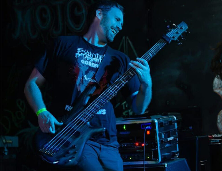 A young man in a black T-shirt plays a bass guitar on stage