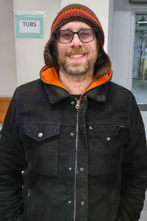 A man stands against a wall wearing a winter coat and hat