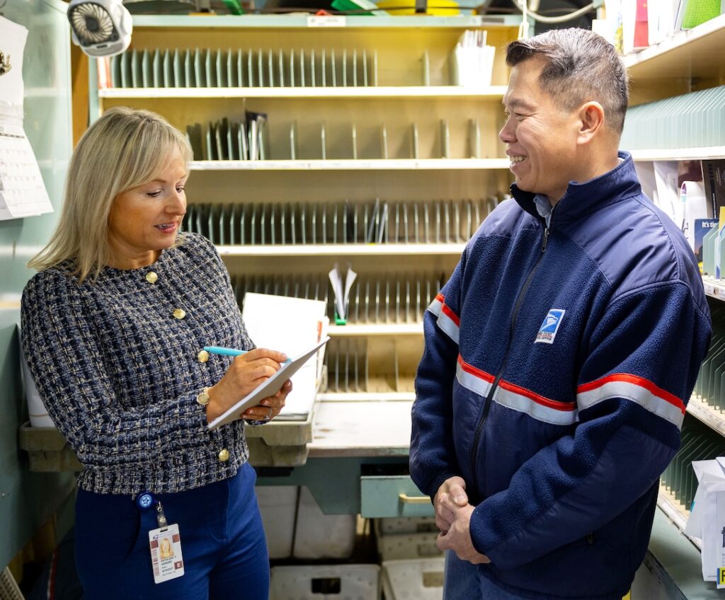 A woman stands in a Post Office workroom and speaks to a uniformed USPS employee while holding a notepad and pen