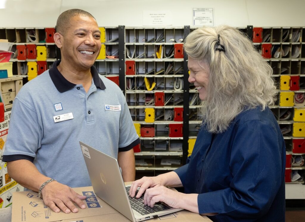 A man in a USPS uniform speaks to a woman as she types on a laptop in a Post Office workroom