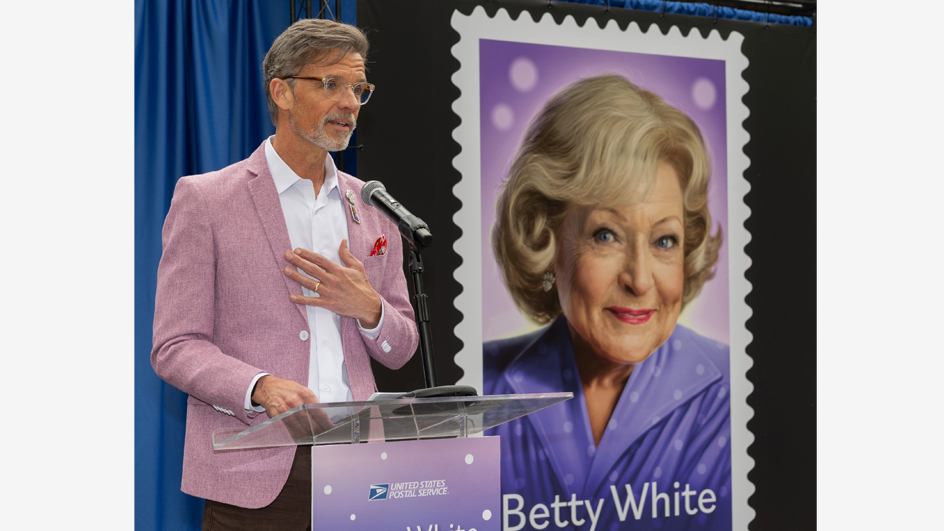 A man in a purple sport coat stands at a lectern and speaks near a poster displaying the Betty White stamp image