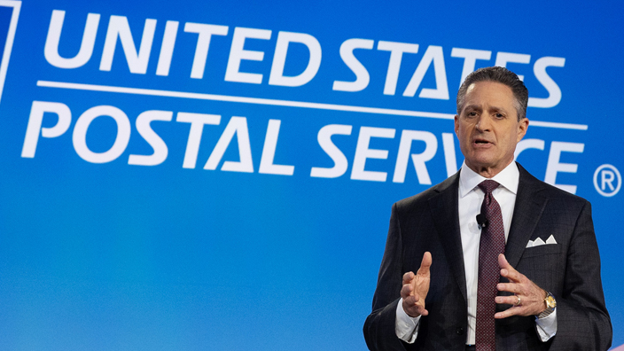 A man in a business suit stands on a stage with the USPS logo looming over his shoulder on a video screen