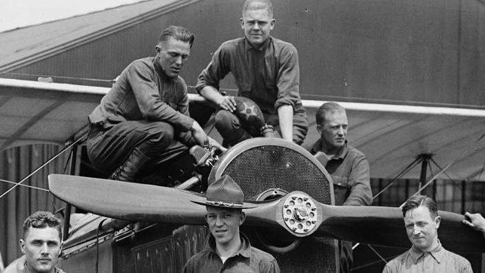 A black and white photo of men in Army uniforms standing near and atop an early 20th-century propeller plane