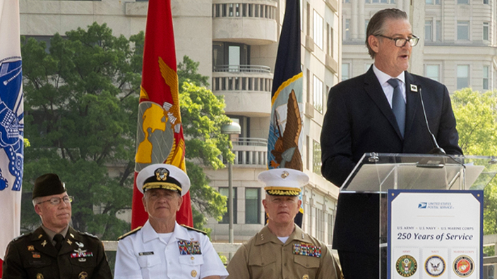 A man in a suit and tie stands at a lectern in front of a large poster bearing military-themed stamp images