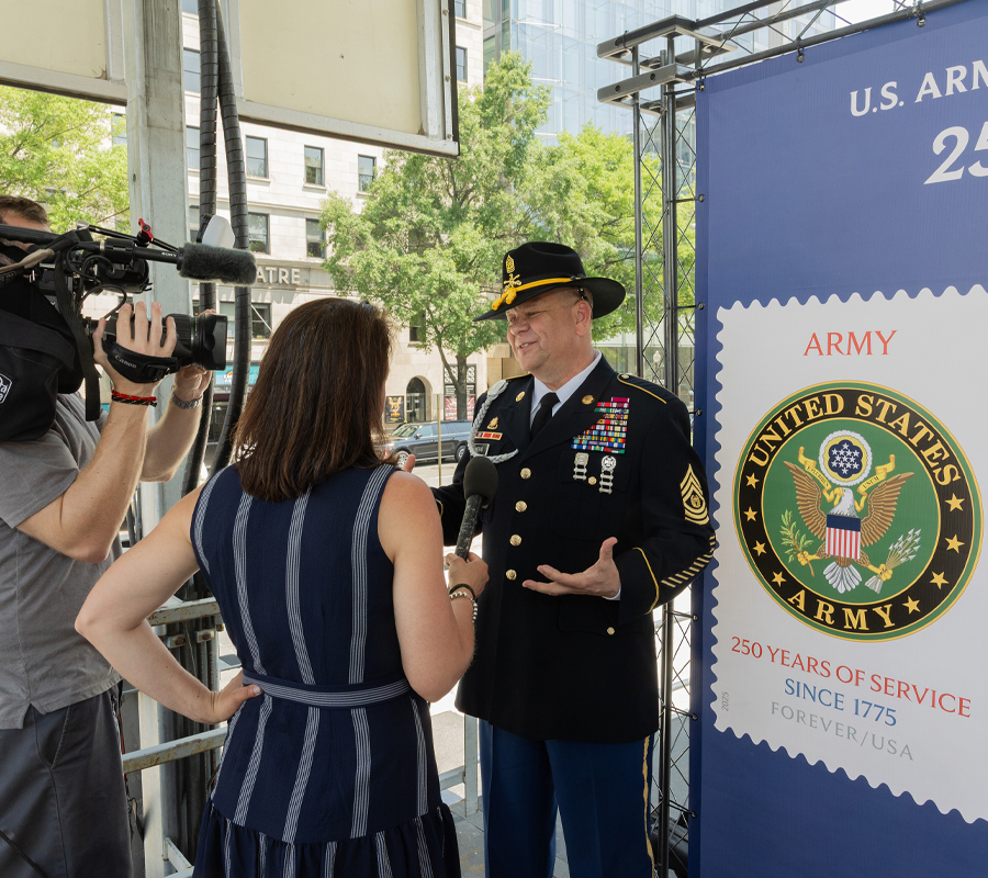 A man in an Army uniform stands in front of a poster bearing military-themed stamp images and addresses a video crew