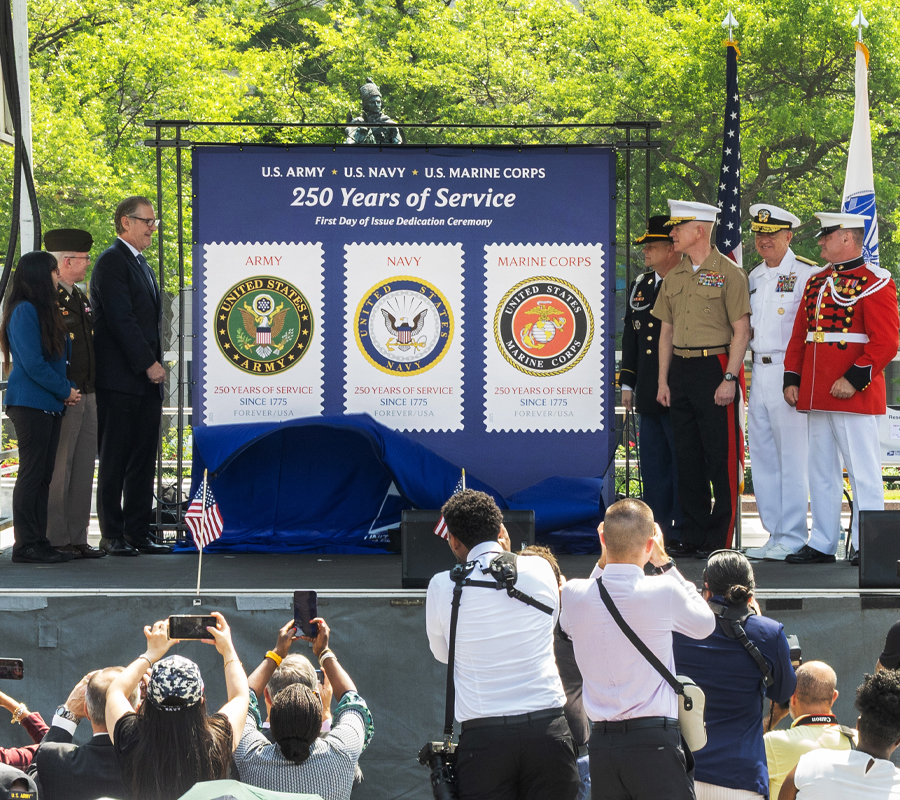 People stand on a stage next to a large poster bearing military-themed stamp images.