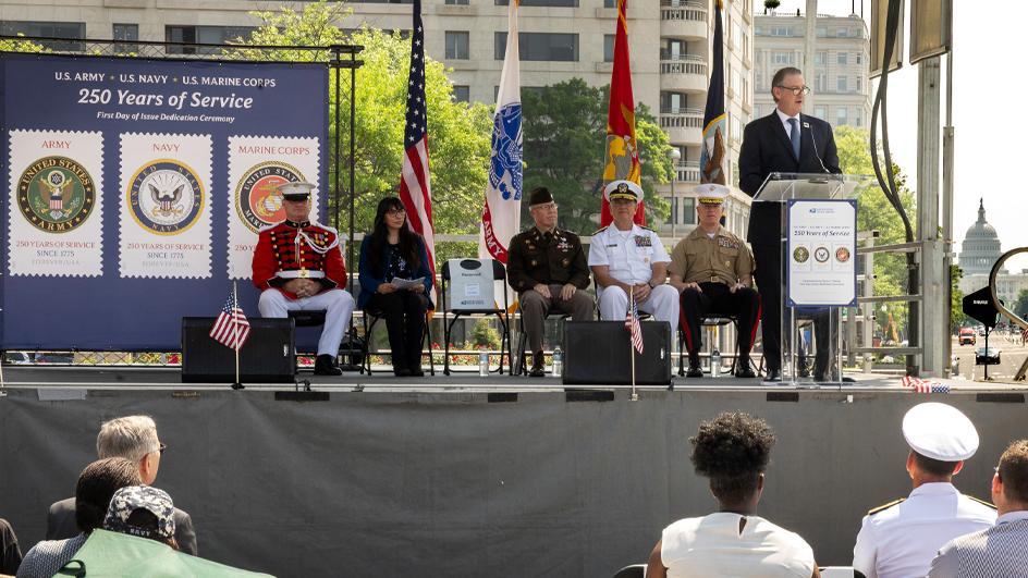 A man in a suit and tie stands at a lectern in front of a large poster bearing military-themed stamp images
