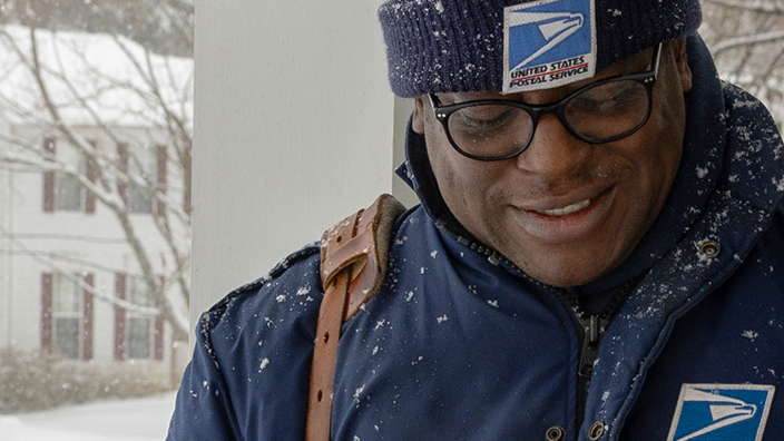 A letter carrier scans a package at a house on a snowy street