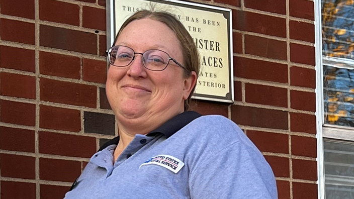 A smiling woman in a Postal Service uniform stands outside a brick Post Office building, holding open the door