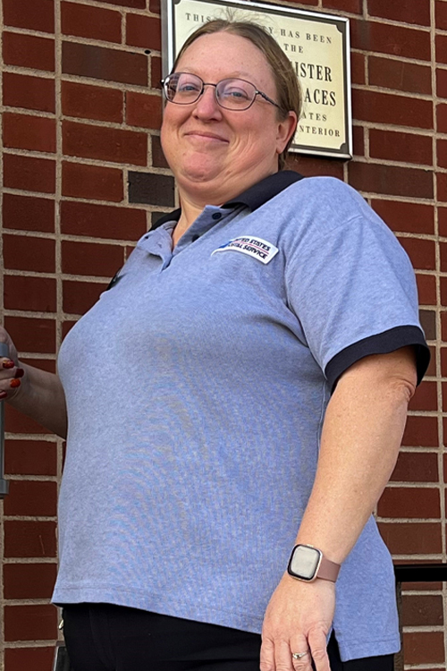 A smiling woman in a Postal Service uniform stands outside a brick Post Office building, holding open the door