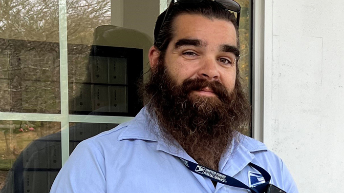 A smiling, bearded man wearing a Postal Service uniform stands outside a building marked as the Pawcatuck, CT, Post Office