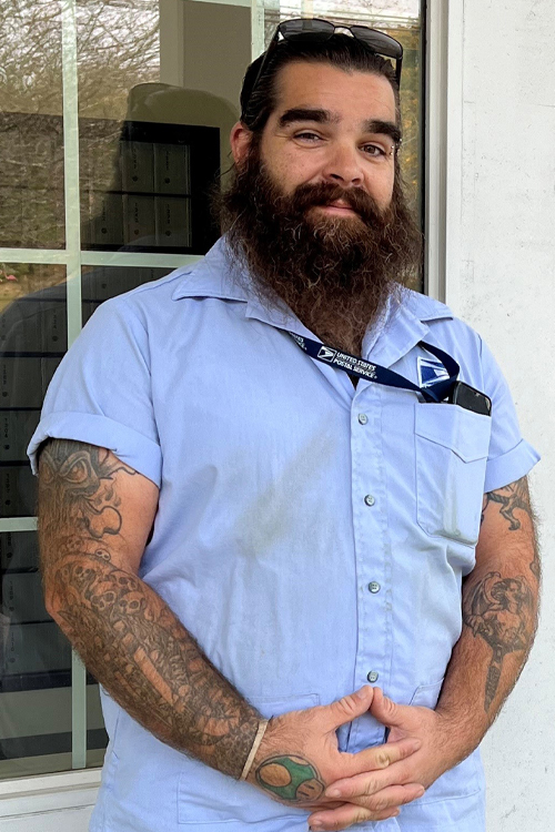 A smiling, bearded man wearing a Postal Service uniform stands outside a building marked as the Pawcatuck, CT, Post Office