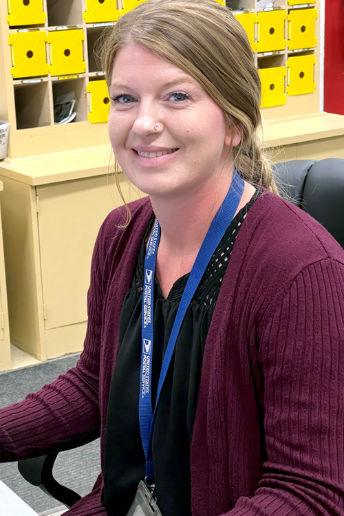 A young woman wearing a cardigan sweater and an ID badge on a lanyard around her neck sits at a desk and smiles
