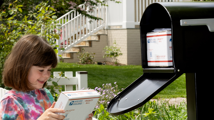 A child stands at a sidewalk mailbox and holds a package