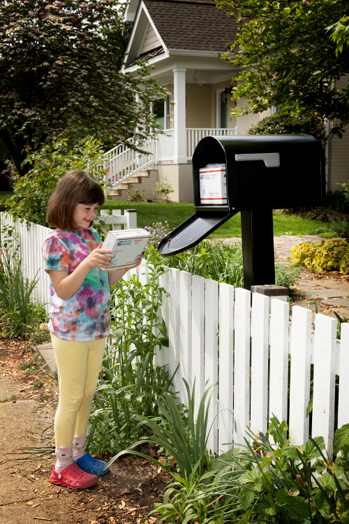 A child stands at a sidewalk mailbox and holds a package