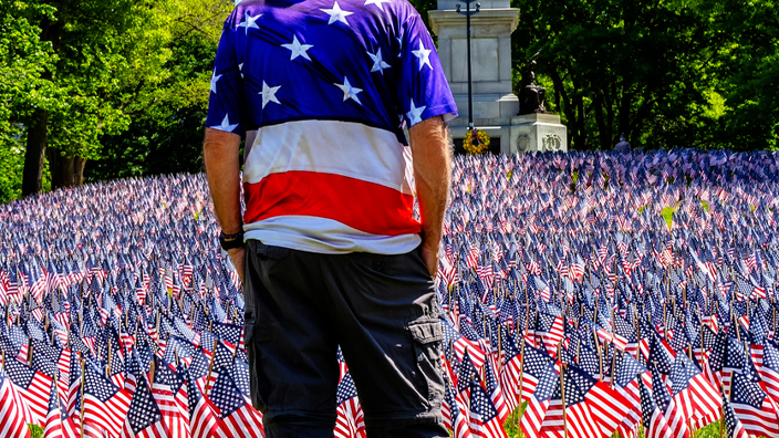 A man wearing a red, white and blue vest — his back to the camera — stands with his head bowed before an array of flags.