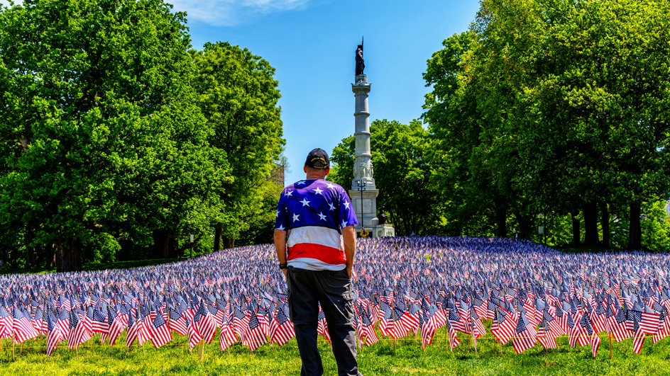 A man wearing a red, white and blue vest — his back to the camera — stands with his head bowed before an array of flags.