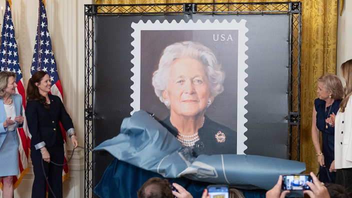 Four women stand on a stage and remove a drape covering a large poster displaying a stamp image of Barbara Bush