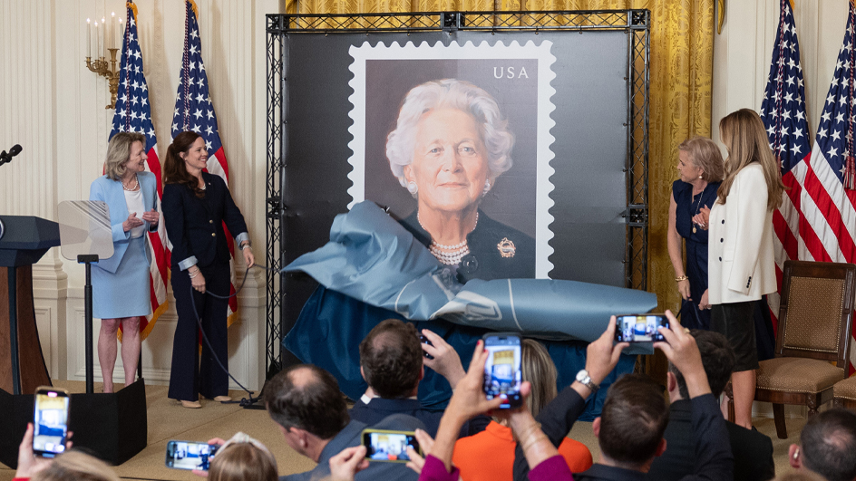 Four women stand on a stage and remove a drape covering a large poster displaying a stamp image of Barbara Bush
