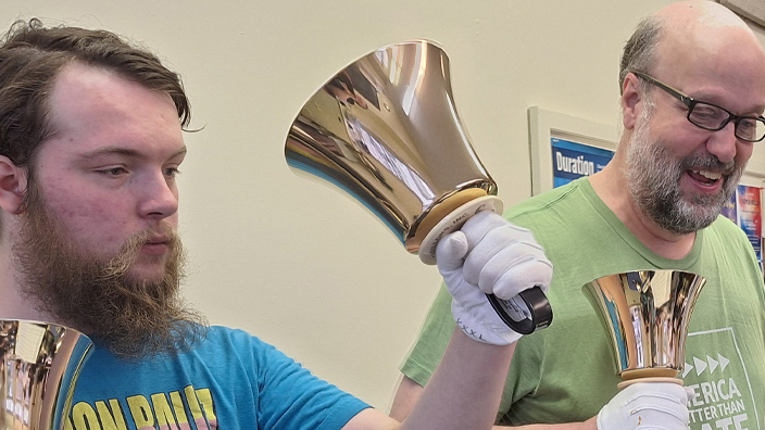 A young man stands next to an older man, each holding large bells in both hands