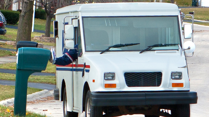 A USPS delivery vehicle stops at a curbside mailbox on a suburban street.