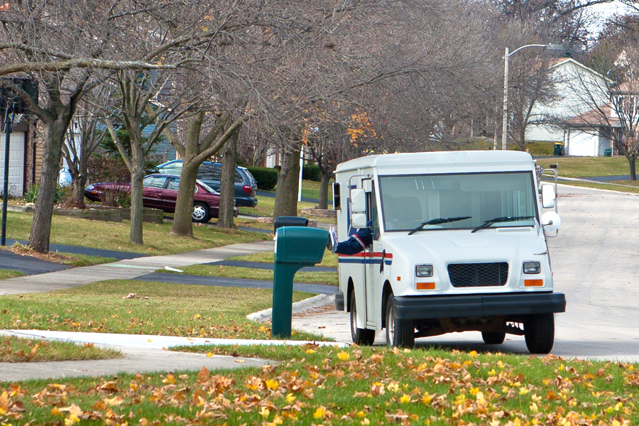 A USPS delivery vehicle stops at a curbside mailbox on a suburban street.
