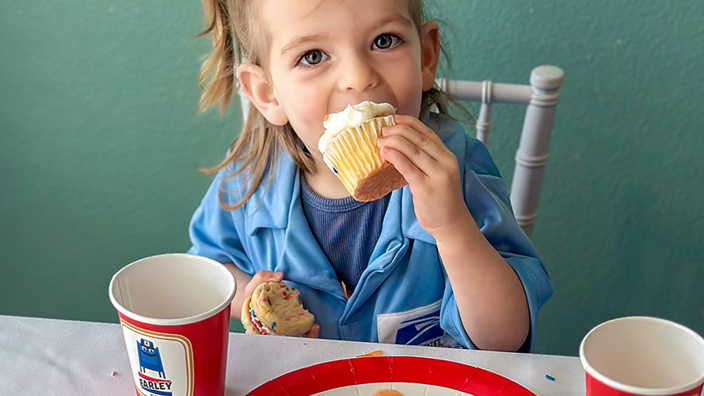 A kid stuffs a cupcake in her mouth while seated near paper plates bearing the image of USPS cartoon characters