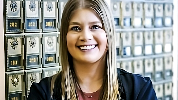 A smiling woman stands with her arms folded near a bank of Post Office boxes