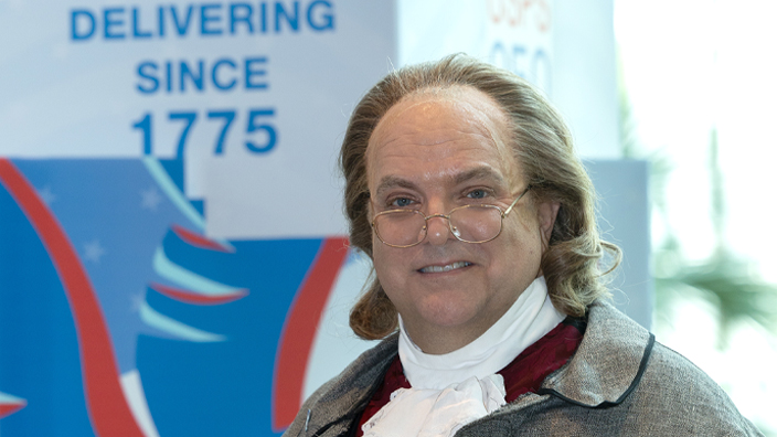 A man dressed as Ben Franklin stands on a convention room floor.