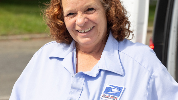 A smiling woman in a postal uniform holds a tray of mail near a delivery vehicle