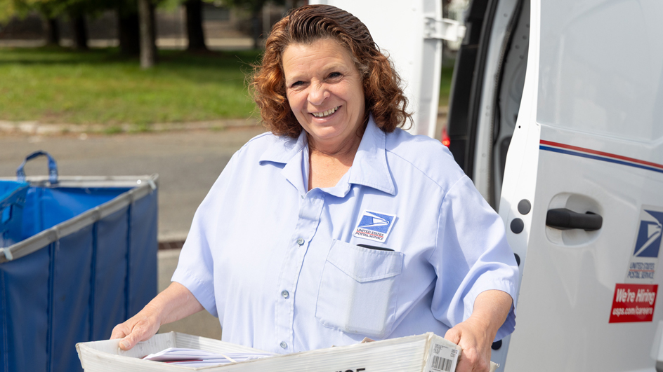 A smiling woman in a postal uniform holds a tray of mail near a delivery vehicle