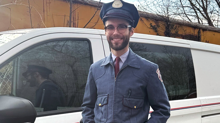 A young man wears an old postal uniform while standing next to a delivery vehicle