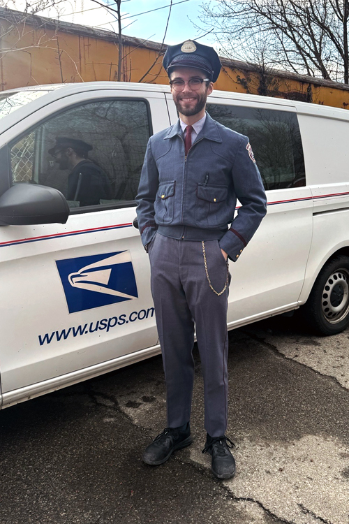 A young man wears an old postal uniform while standing next to a delivery vehicle