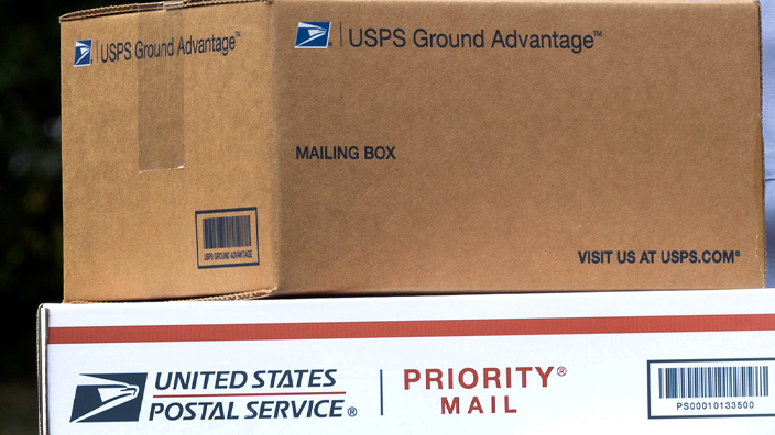 A letter carrier, seen from the neck down, holds USPS-branded packages