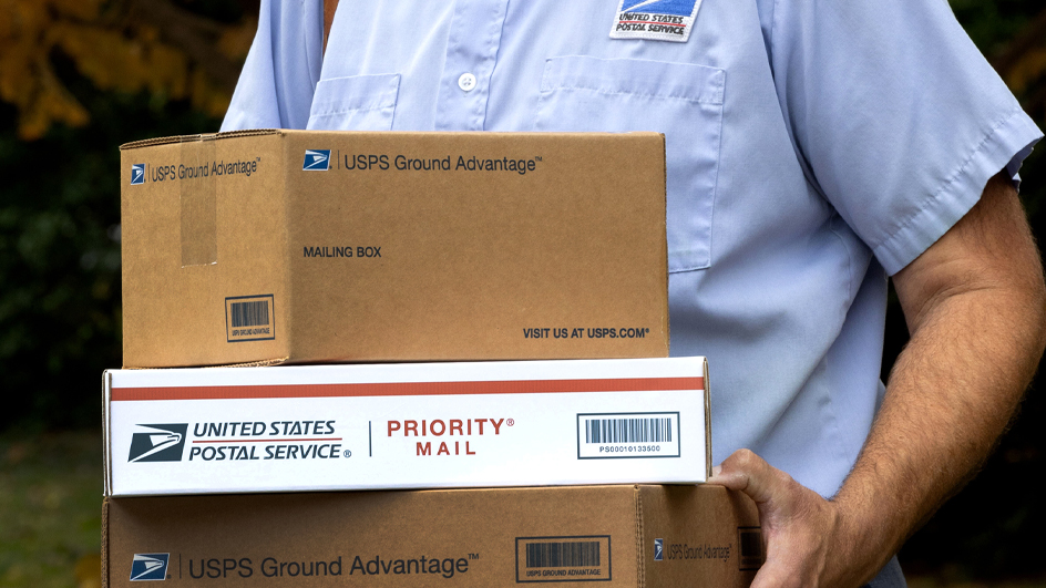 A letter carrier, seen from the neck down, holds USPS-branded packages