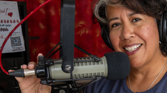 A smiling woman sits at a console and holds a microphone inside a radio studio