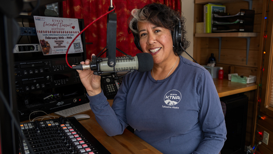 A smiling woman sits at a console and holds a microphone inside a radio studio
