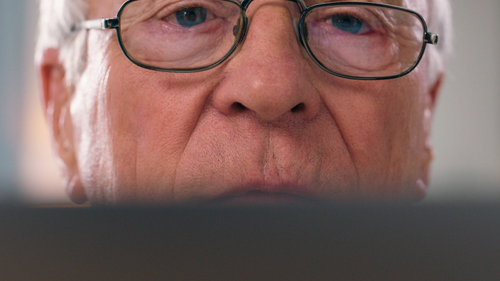 An older man peers over a computer screen with a skeptical expression on his face