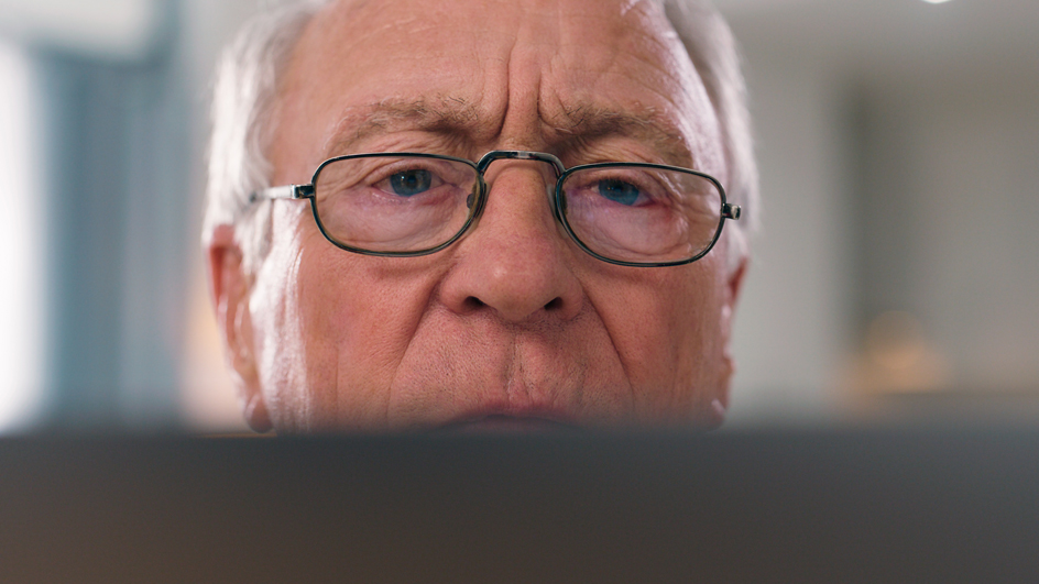 An older man peers over a computer screen with a skeptical expression on his face