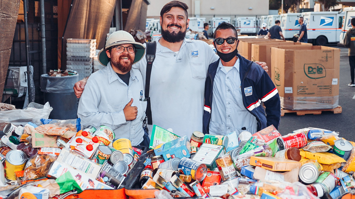Three men in postal uniforms stand near a huge bin filled with canned and boxed food items