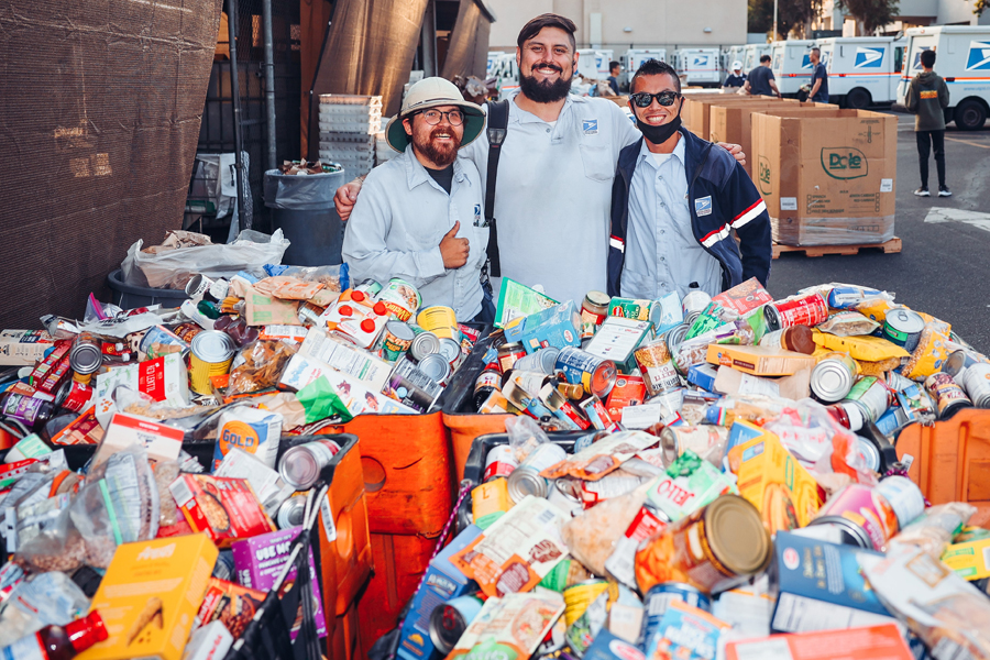 Three men in postal uniforms stand near a huge bin filled with canned and boxed food items