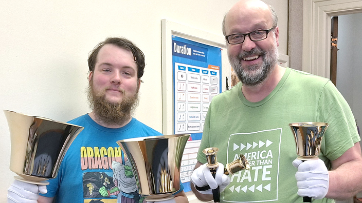 A younger man and an older man wear T-shirts and stand in a hallway holding large bells