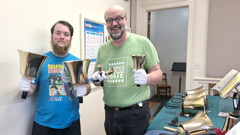 A younger man and an older man wear T-shirts and stand in a hallway holding large bells