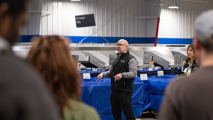 A man stands on the floor of a postal processing plant, addressing a group of workers