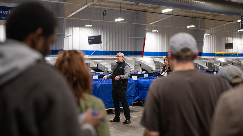 A man stands on the floor of a postal processing plant, addressing a group of workers